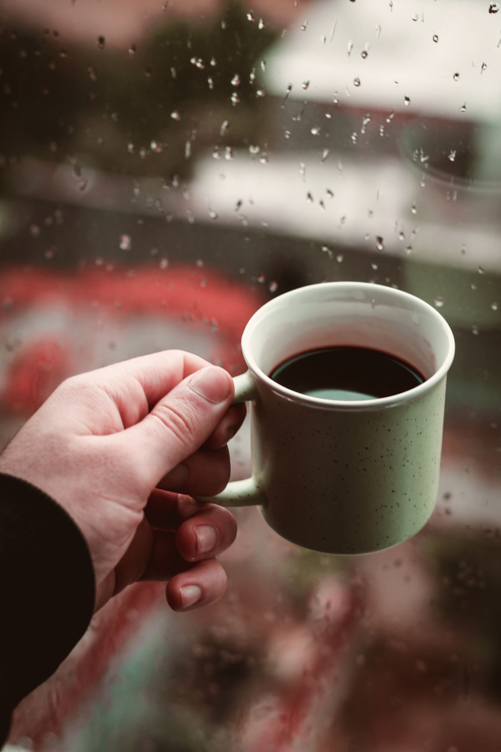 Mão segurando uma xícara de café junto a uma janela com chuva, clima de tensão contida